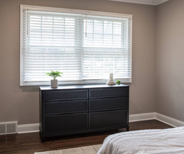 Black dresser below a window with blinds, topped with small plants and decor in a minimalist bedroom.