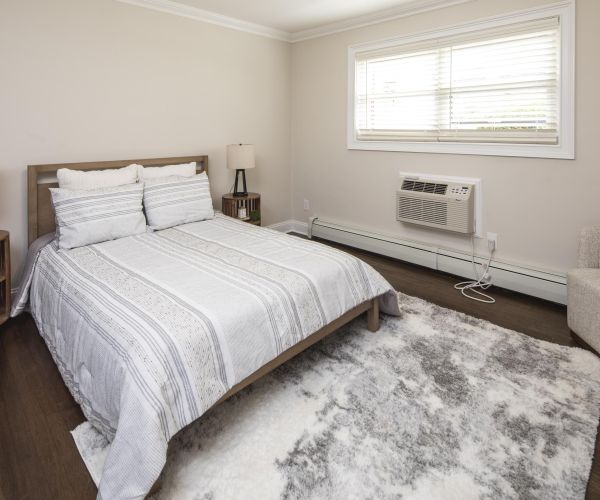 A cozy bedroom with a neatly made bed, two bedside tables, a window, and an armchair on a fluffy rug.
