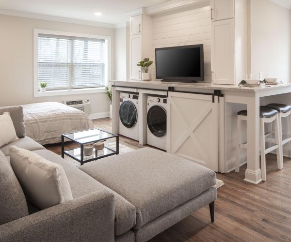 Modern studio apartment with grey sofa, bed, kitchen island, and washer-dryer under a bar counter.