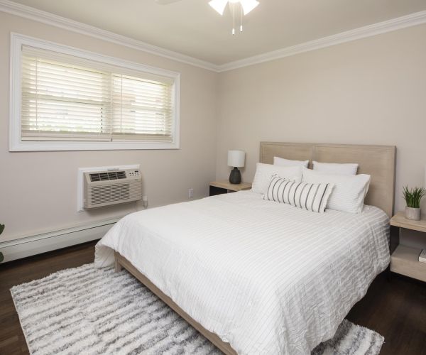 A neatly made bed in a bright, modern bedroom with neutral décor and a window air conditioner.