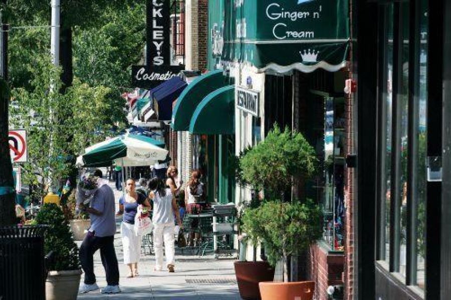 People stroll along a sunny Englewood sidewalk lined with shops, cafes, and green awnings on a busy street.