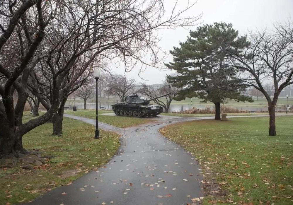 A military tank sits on grass near a path in a Clifton park with bare trees and overcast sky.