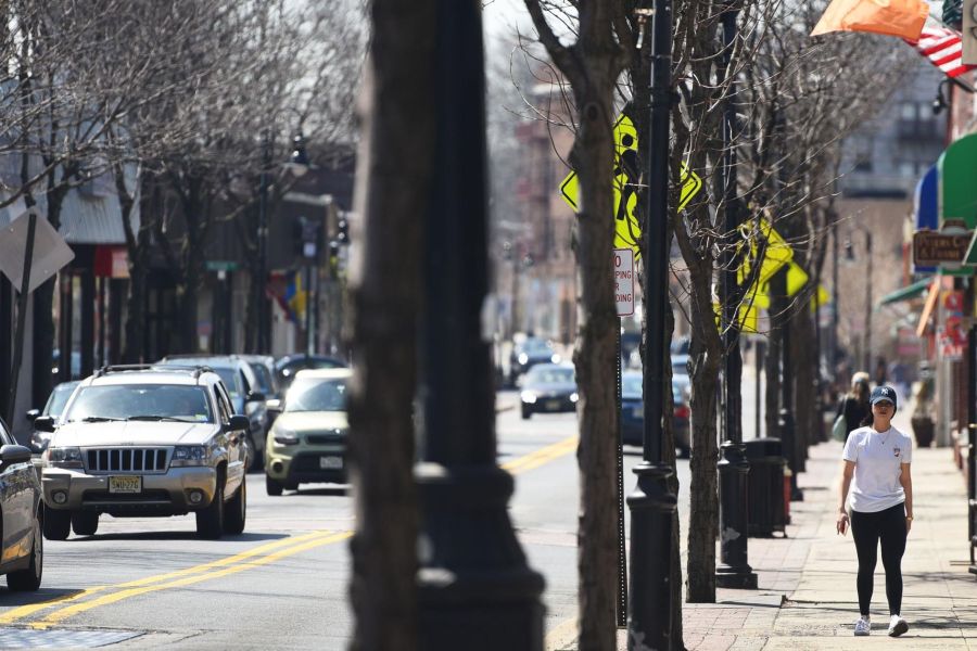 A woman walks on a Bergenfield city sidewalk beside parked cars and traffic on a sunny day.