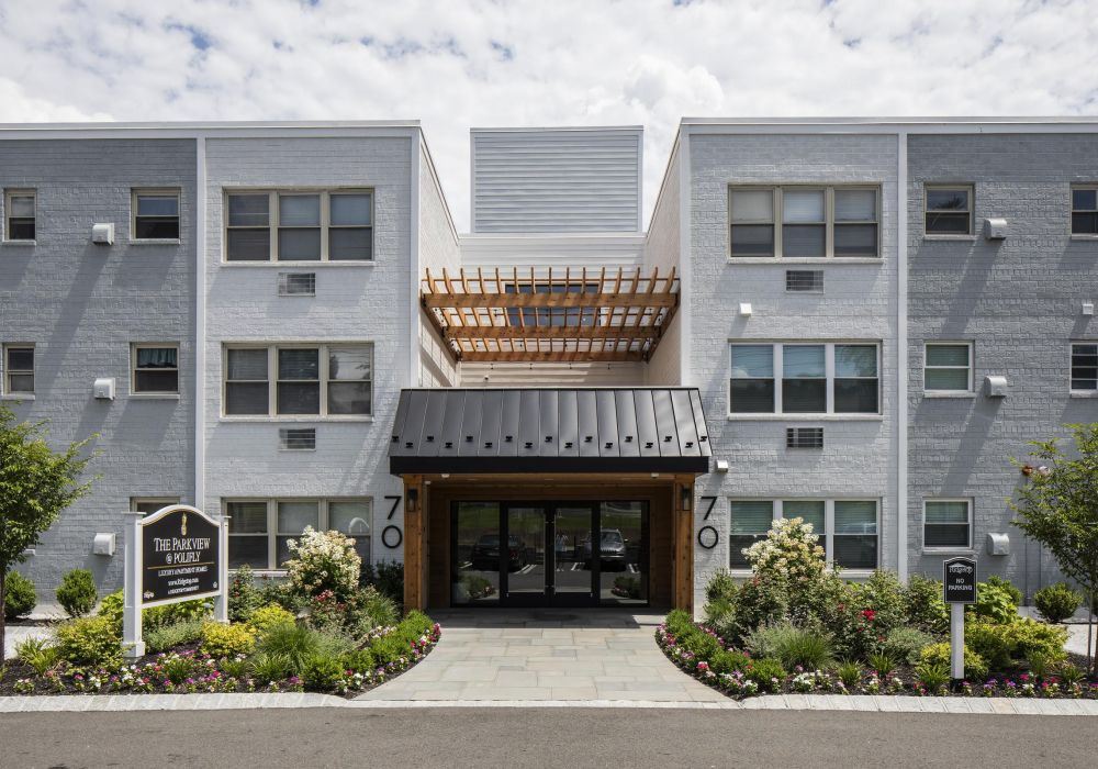 Modern three-story apartment building with a landscaped entrance and a wooden pergola over the doorway.