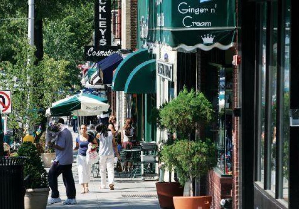 People stroll along a sunny Englewood sidewalk lined with shops, cafes, and green awnings on a busy street.